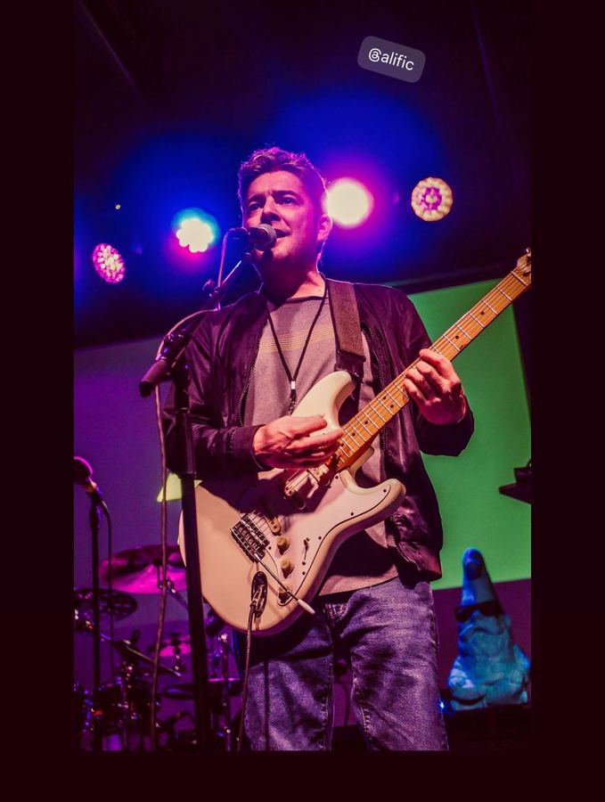 Alific singing and playing a white Stratocaster under purple and blue stage lights