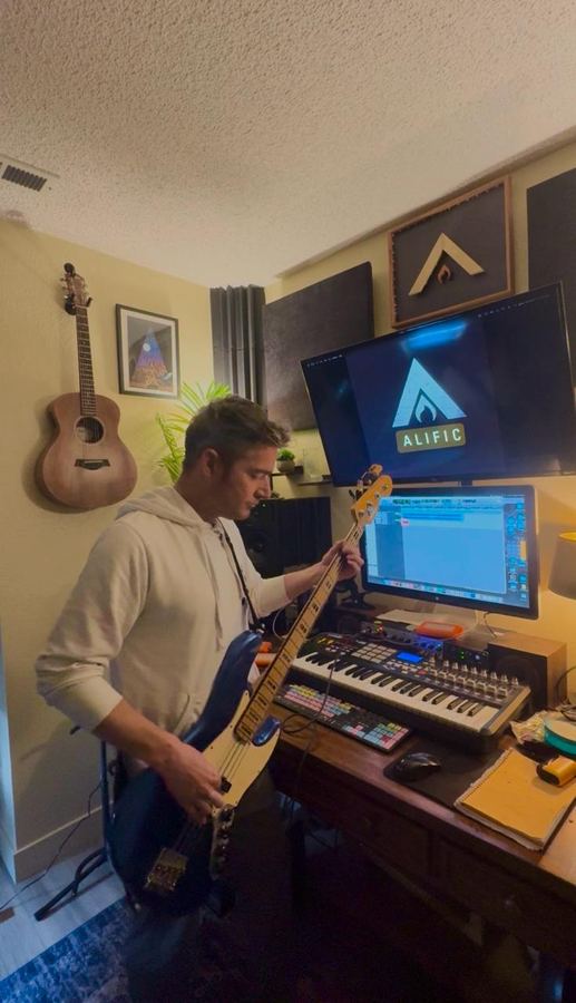 Alific playing a blue bass guitar in his studio, the Alific logo on the screen behind him