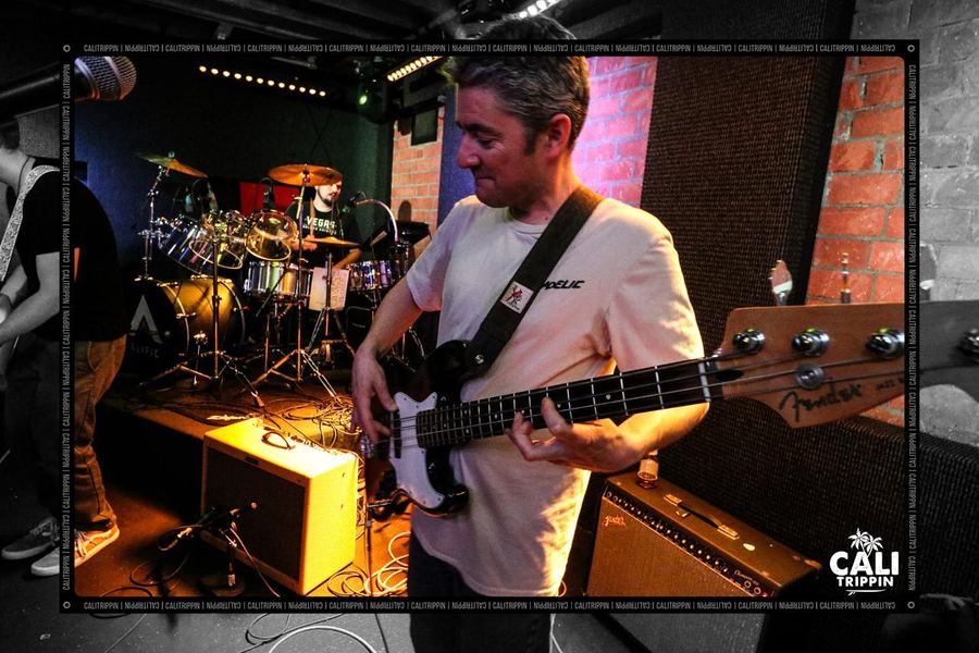 Alific playing a white Fender bass on stage in front of a brick wall, drum kit behind him