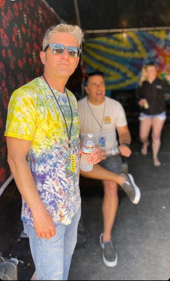 Alific backstage in a tie-dye shirt and sunglasses with an artist pass around his neck