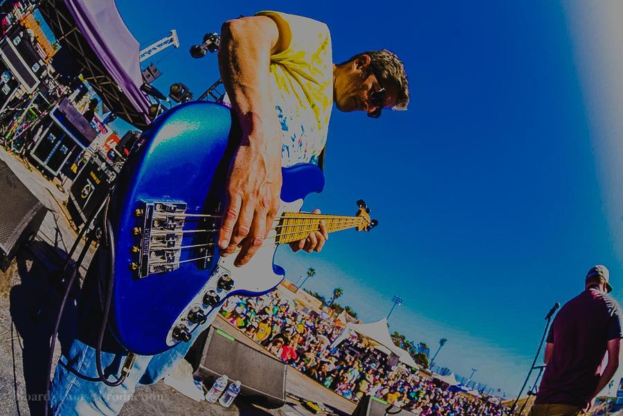 Alific playing a blue bass in a fisheye view of a packed festival stage, the crowd stretched out below him