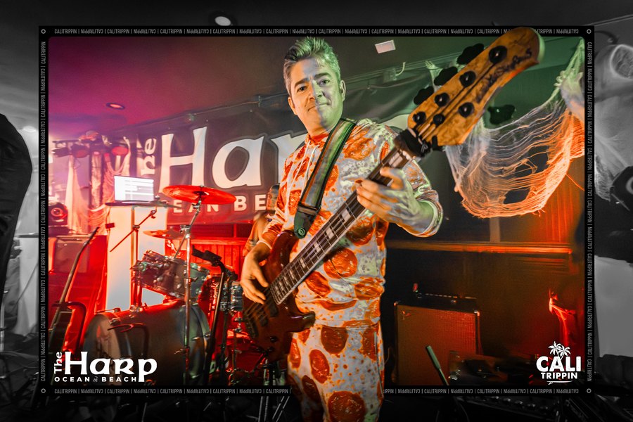 Alific performing on bass at The Harp Ocean Beach in a Halloween pumpkin suit, band behind him on a lit stage